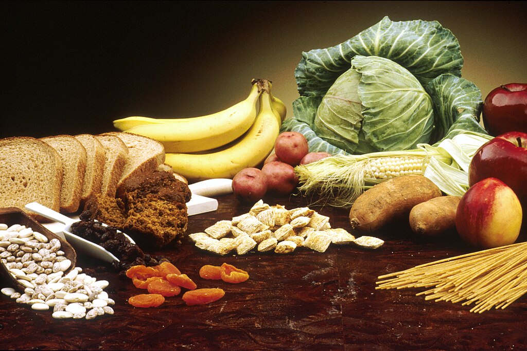 A wooden table containing: a ladle full of beans, a sliced loaf of brown bread, a bunch of bananas, muffins, small potatoes, a head of cabbage, an ear of corn, a pile of cereal, yams, apples, a nectarine and some spaghetti. Source: https://commons.wikimedia.org/wiki/File:Fruit,_Vegetables_and_Grain_NCI_Visuals_Online.jpg